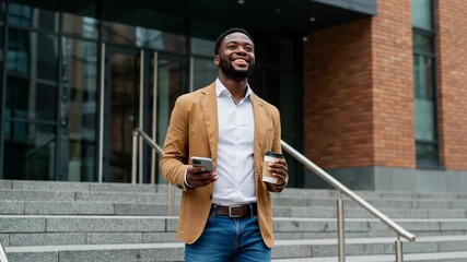 A smiling black man in a tan blazer and jeans walks down steps outside a modern office building, holding a phone and a coffee cup - Powered by Adobe