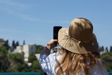 Back view of a woman wearing a wide brimmed hat, standing outdoors and photographing nature with a...