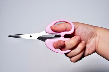 Hand holding closed small paper scissors with pink colored handle isolated on horizontal gray background.