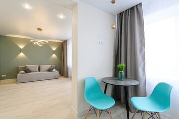 bright room features teal chairs, a round table, and grey curtains by a window. A glimpse into a...