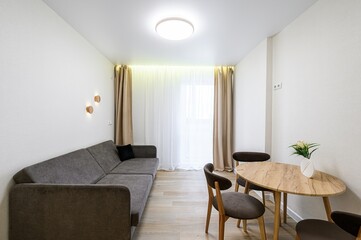 Bright living room with a grey sofa, round wooden table, and chairs. Sheer curtains and beige...