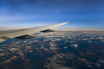 View through an airplane window catching the amazing mountains through  the clouds, with the plane's wing prominently displayed against the horizon.
