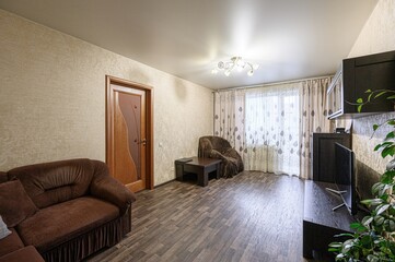 cozy living room with brown furniture, a wooden floor, and light-colored walls. A window with...