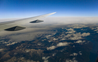 View through an airplane window catching the amazing mountains through  the clouds, with the plane's wing prominently displayed against the horizon.