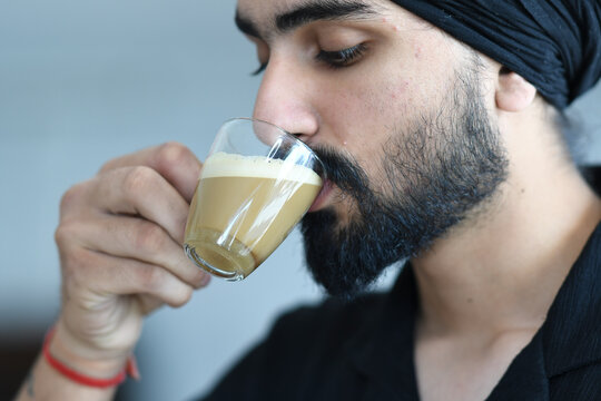 A bearded man with a black turban drinks from a clear glass filled with a creamy beverage, enjoying a moment of relaxation and savoring the taste of his drink with closed eyes in a close-up shot - Powered by Adobe