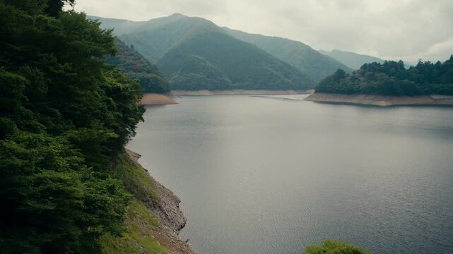 Wide Oku Tama Lake framed by lush green trees and misty mountain horizon &ndash; Scenic cloudy day capturing calm Japanese lake environment.
