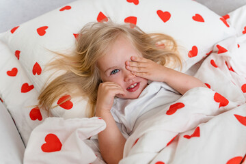 Little Caucasian girl child in bed under a white blanket with hearts smiling