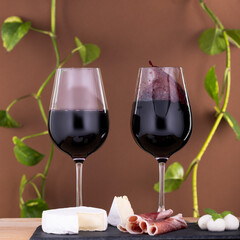 Two glasses of red wine with cheese and prosciutto on slate board against brown background. Perfect for concepts of fine dining, celebration, and Mediterranean lifestyle.