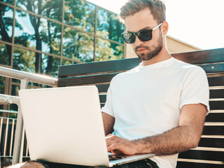 Handsome smiling stylish hipster lambersexual model. Fashion man sitting on the bench in the street...
