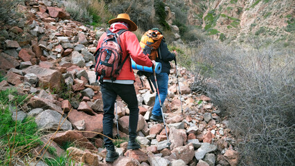 Tourists with backpacks and trekking poles walk along a rocky path. Two people climb a dangerous...