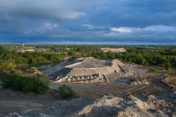 Open-pit mining or quarry landscape with soil heaps, green trees and dramatic cloudy sky, symbol of industry, environment and land use.