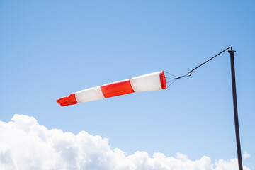 Red-white striped windsock against a clear blue sky with fluffy white clouds in summer as an...