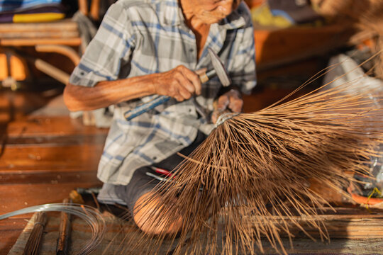 Elderly Thai craftsman hammering coconut leaf broom at dusk