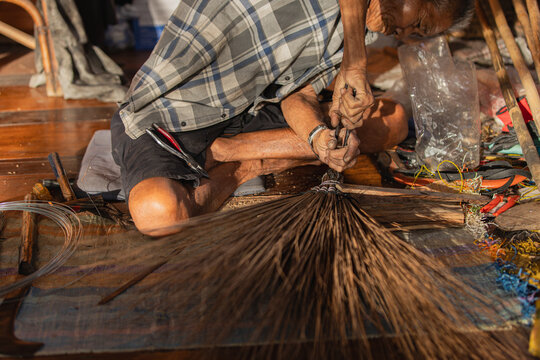 Elderly Thai artisan binding coconut leaf broom at dusk