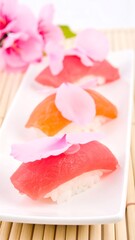 Three pieces of sushi, adorned with pink petals, rest on a white rectangular plate atop a bamboo mat, with pink flowers in the background