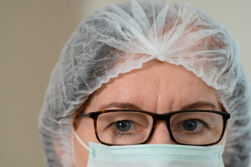 Close-up portrait of a mature female doctor wearing a medical mask and surgical cap. Her calm, focused gaze reflects professionalism, health care commitment, and patient safety.