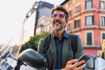 Confident middle-aged man in a blazer with mobile phone and backpack, smiling beside scooter in the...