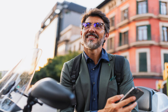 Confident middle-aged man in a blazer with mobile phone and backpack, smiling beside scooter in the city. Concept of freedom, connection, modern business lifestyle