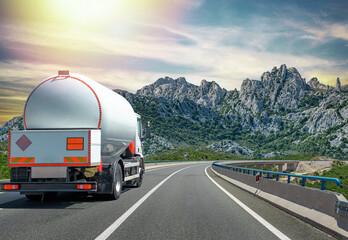 A large tanker truck transporting fuel drives along a curved highway through a mountainous landscape under a bright sky.