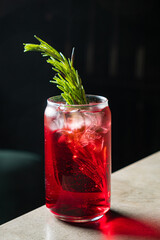 Bright red drink with ice and a fragrant herb garnish sits elegantly on a textured surface