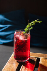A vibrant red beverage garnished with fresh rosemary sits on a rustic wooden table in soft lighting