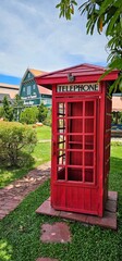 Fototapeta premium one big red telephone booth stands in the yard of a house on the street, there is a green lawn and grass with green trees. in the background there is a house with an orange tiled roof. there are white