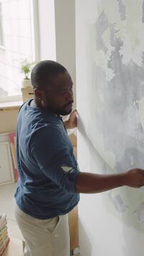 Vertical shot from above of African American man in blue shirt scraping off old paint from wall while carrying out renovation in living room