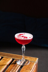 Deep red cocktail with a frothy top and sprinkled garnish rests on a rustic wooden table