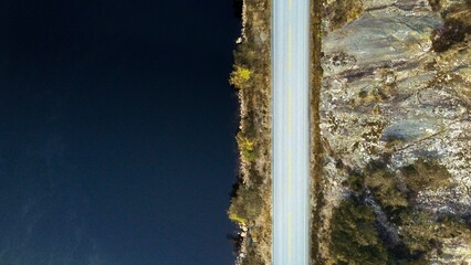Top down view of a beautiful road in Norway (Mykland) next to a dark blue lake. Rocks and a few trees on the right and left side of it.