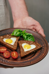 person holds clay plate with two pieces brown bread shaped into triangles, each with sunny side up egg. cherry tomatoes and fresh basil accompany dish. vertical. close up.