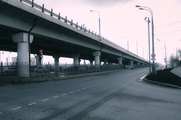 Modern city transport interchange with a high overpass bridge, supports and street lamps. Empty asphalt road goes into the distance under a gray sky. Cool atmosphere, urban, with a feeling of silence 