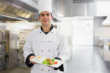Male chef in chef coat holding plate topped with fish fillet, greens, ramekin in restaurant kitchen