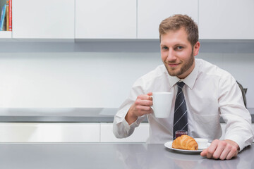 Businessman sitting at countertop in kitchen holding coffee mug and croissant with jam, copy space