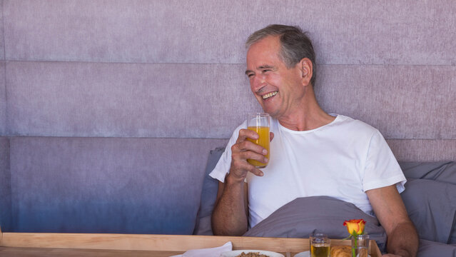 Senior man wearing white tee sitting on bed drinking orange juice near breakfast tray, copy space