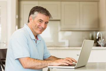 Senior man typing on silver laptop in modern kitchen at table with marble countertop