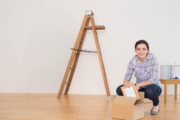 Woman kneeling in room unpacking frame from cardboard box beside ladder and paint cans, copy space