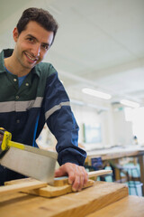 Man wearing reflective coverall sawing wooden plank with yellow-handled saw at workshop, copy space