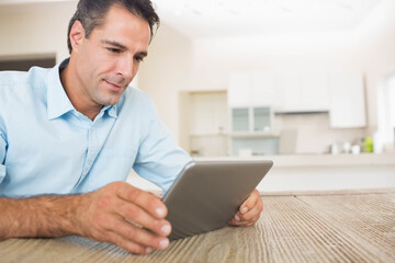 Man wearing light-blue shirt leaning on wooden table using tablet in bright kitchen, copy space