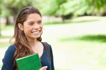 Smiling woman student holding green book and wearing backpack while standing in park, copy space