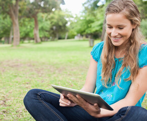 Fototapeta premium Woman sitting cross-legged on grass in public park holding tablet and smiling, copy space