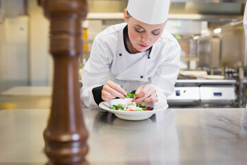 Female chef leaning over counter in commercial kitchen placing fresh herbs on salad bowl