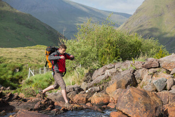 Adult male hiker wearing jacket carrying backpack stepping across stream in valley, copy space