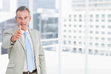 Mature male standing in office before glass windows wearing suit giving thumbs up, copy space