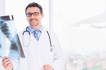 Male physician reviewing chest X-ray film at window in clinic wearing lab coat and stethoscope