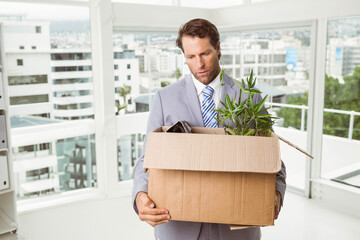 Middle-aged man in suit standing in skyline office, holding cardboard box with plant and binder