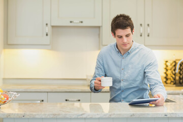 Man standing behind marble countertop in home kitchen holding coffee mug, using tablet, copy space