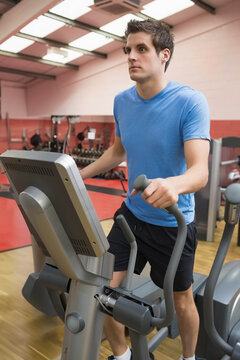 Fototapeta Male wearing blue shirt working out on elliptical trainer in gym amid dumbbells and cardio machines