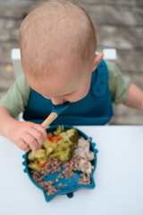 Overhead view of a toddler eating from a bowl with a spoon in a nature setting. First meal with...