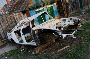 Severely damaged, rusted white car abandoned in grassy area, propped on timber. Heavy corrosion...