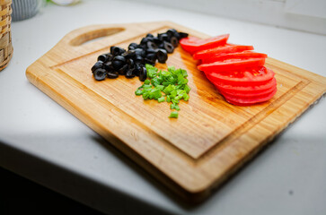 A rustic wooden cutting board with black olives, chopped green onions, and tomato slices on a white countertop, perfect for salad or Mediterranean dish preparation.